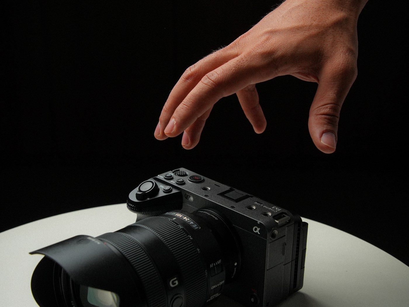 Mão alcançando a câmera Sony FX3 sobre a mesa, sob luz lateral, em fundo escuro